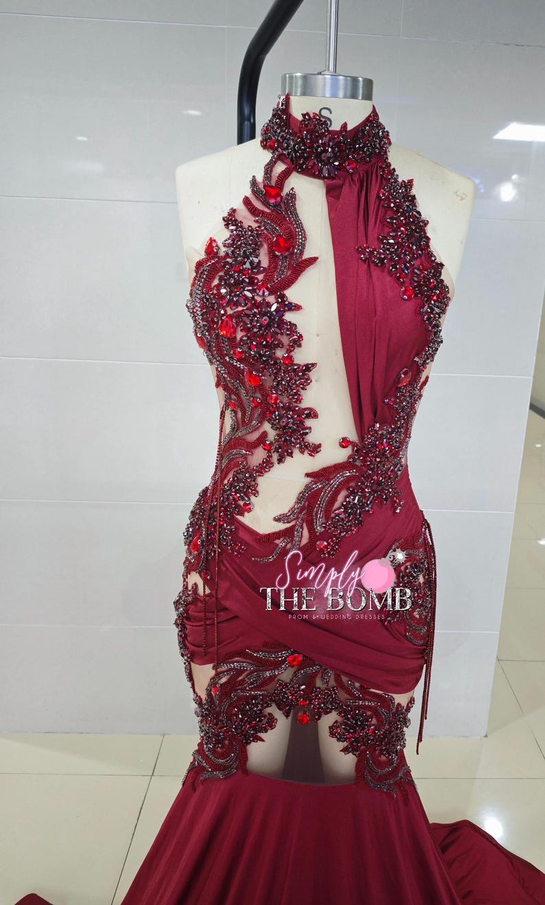 May include: A burgundy halter-neck dress with intricate red beading and embellishments. The dress features a fitted bodice, a sheer panel, and a draped skirt. The text "Simply The Bomb" is visible.