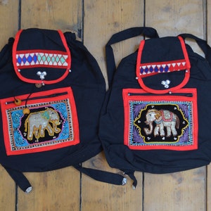 May include: Two black backpacks with red trim and embroidered elephant designs. Each bag has a flap with colourful geometric patterns and a front pocket with a zip. The bags are made of a soft fabric.