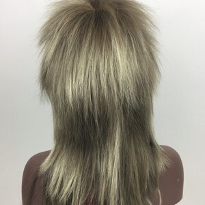 Op de afbeelding: Een lange, gelaagde pruik in een lichtbruine en blonde kleur. De pruik is gestyled met een middenscheiding en heeft een ruige, textuurrijke look.