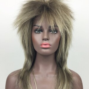 Op de afbeelding: Een lange, gelaagde pruik in een lichtbruine en blonde kleur. De pruik heeft een stekelige, gevederde stijl.