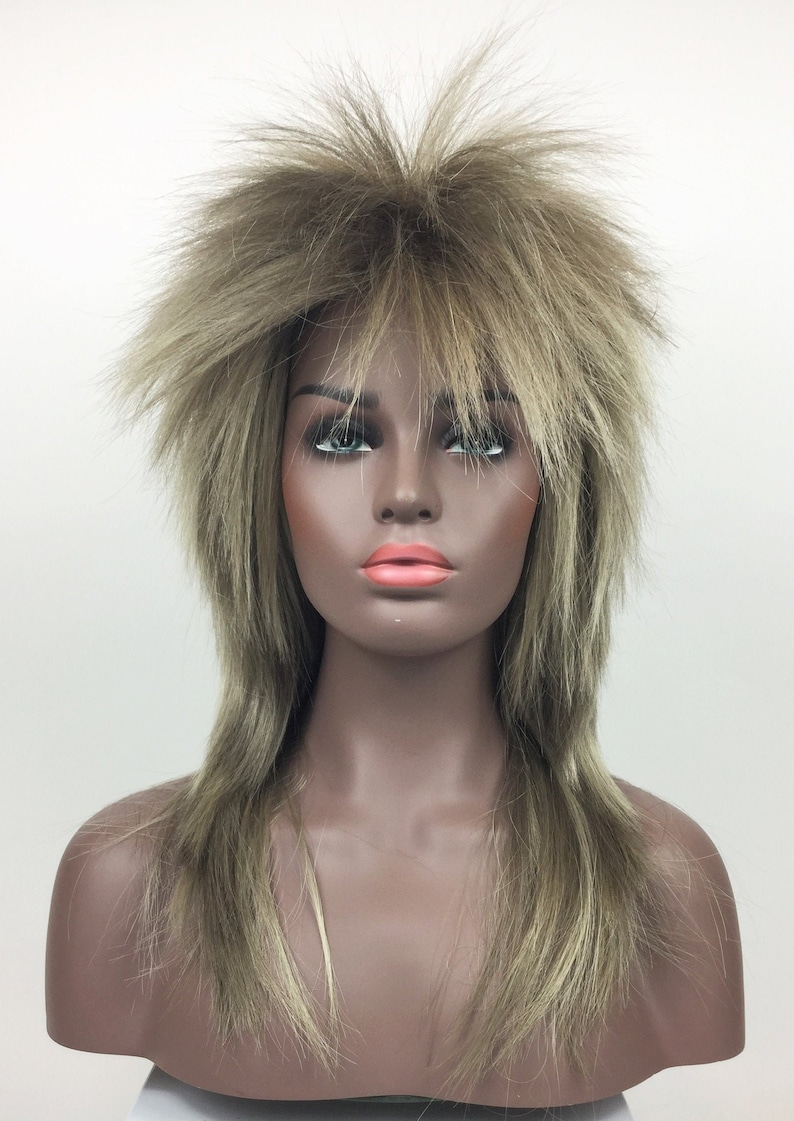 Op de afbeelding: Een lange, gelaagde pruik in een lichtbruine en blonde kleur. De pruik heeft een stekelige, gevederde stijl.