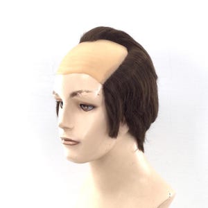 May include: A brown hairpiece with a light skin-toned cap, designed to simulate a receding hairline. The hair is styled with short, side-swept bangs and a slightly longer back. The hairpiece is displayed on a mannequin head.