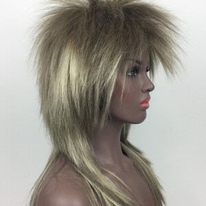 Op de afbeelding: Een lange, gelaagde pruik met een stekelige, stompe stijl. De pruik is lichtbruin met blonde highlights.