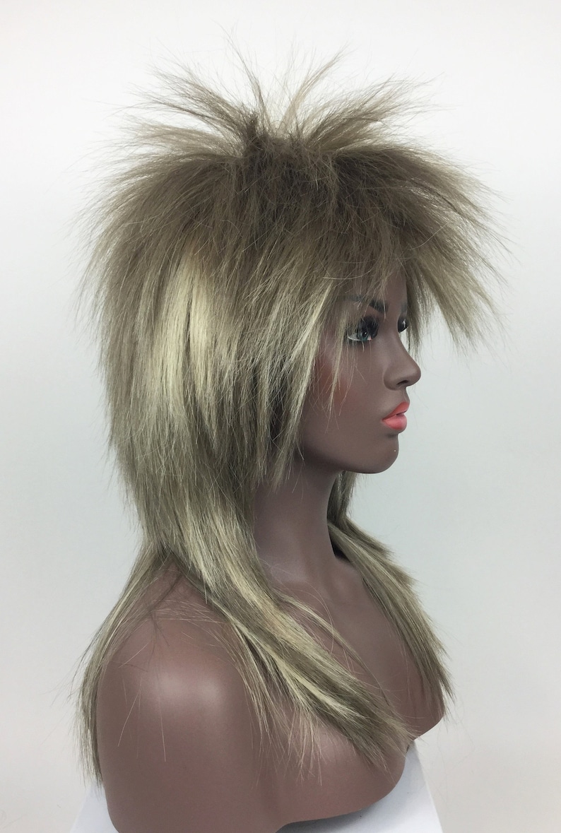 Op de afbeelding: Een lange, gelaagde pruik met een stekelige, stompe stijl. De pruik is lichtbruin met blonde highlights.