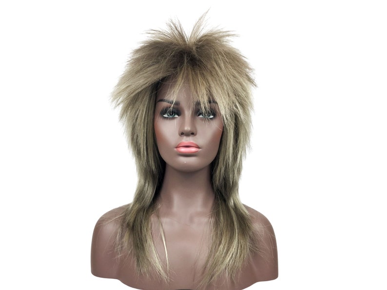 Op de afbeelding: Een lange, gelaagde pruik met een ruige, stompe stijl. De pruik is lichtbruin met blonde highlights.