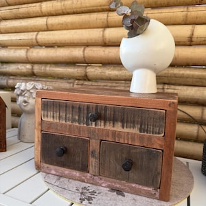 Könnte beinhalten: Eine kleine, rustikale Holzkiste mit vier Schubladen und schwarzen Knöpfen. Die Kiste besteht aus Altholz in Braun- und Grautönen. Eine weiße, kugelförmige Vase mit kurzem Stiel hält einen Zweig mit Grün. Die Kiste steht auf einer runden, gemusterten Matte.