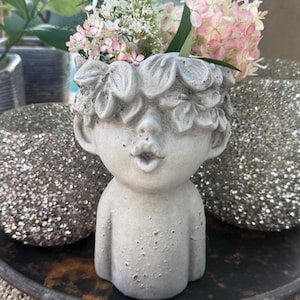 Könnte beinhalten: Ein grauer Betonblumentopf in Form eines Kindergesichts, mit einem Blumenkranz und offenem Mund. Er ist mit rosa und weißen Blumen und Grünzeug gefüllt. Der Topf steht auf einem dunklen Metalltablett.