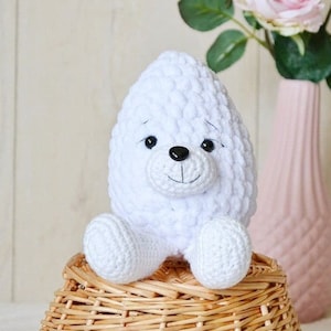 Op de afbeelding: Wit gehaakt knuffeldier met zwarte ogen en een zwarte neus, zittend op een rieten mand. Het dier heeft de vorm van een beer of een hond.