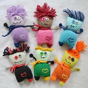 May include: Six small, handmade crochet dolls featuring yarn hair, button eyes, and embroidered heart details on their torsos. Each doll displays a unique color combination, including purple, brown and pink, light blue, black and white, green, and orange.