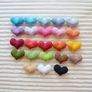 May include: Twenty-four crocheted hearts in a variety of colours, including red, pink, blue, green, yellow, orange, brown, and black. The hearts are arranged in rows on a white textured surface.