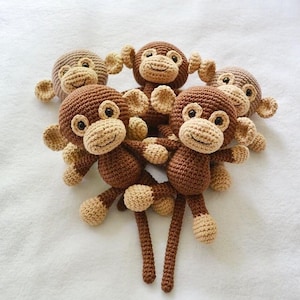 Op de afbeelding: Een verzameling van vijf kleine, handgemaakte gehaakte apen. Elke aap heeft een donkerbruin lichaam, een lichter beige gezicht, oren, handen en voeten, met zwarte geborduurde ogen en een kleine glimlach. De apen zijn in een groepje gerangschikt.