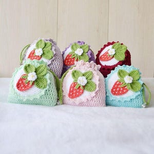 May include: A collection of six crocheted drawstring bags in various pastel colors. Each bag features a white circular appliqué with a red strawberry, green leaves, and a small white flower. The bags are handmade and have a delicate, feminine aesthetic.