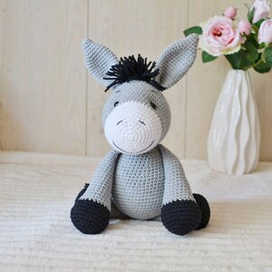 Pode incluir: Um burro de peluche cinza crochetado com cascos pretos e uma crina preta. O burro está sentado em uma superfície branca e fofa.