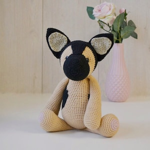 May include: A crocheted stuffed dog toy with black and tan fur, sitting on a white surface. The dog has floppy ears and a black nose.