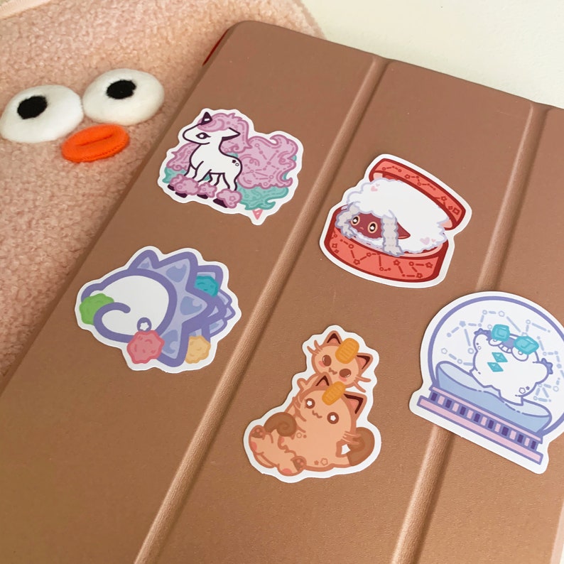 Puede incluir: Cinco pegatinas de personajes de dibujos animados de colores sobre una superficie marr&oacute;n. Las pegatinas representan un unicornio rosa, una oveja blanca en una caja roja, un dinosaurio azul y morado, un gato marr&oacute;n y un gato blanco en una bola de nieve.