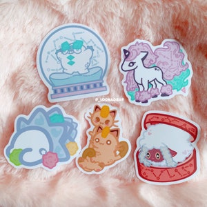 Puede incluir: Cinco pegatinas coloridas con personajes de dibujos animados de la franquicia Pok&eacute;mon. Las pegatinas est&aacute;n sobre un fondo rosa peludo. Las pegatinas representan un Pok&eacute;mon azul y blanco en una bola de nieve, un Pok&eacute;mon unicornio rosa y blanco, un Pok&eacute;mon marr&oacute;n y naranja, un Pok&eacute;mon azul y blanco y un Pok&eacute;mon rojo y blanco en una caja.