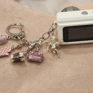 May include: A white digital camera with a black screen, attached to a keychain with charms. Charms include a pink baseball cap, oven mitt, coffee cup, purse, and a silver angel. The camera and charms are on a pink surface.