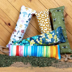May include: Five colorful catnip toys in the shape of long pillows. The toys are made of fabric with different patterns, including mushrooms, bees, and abstract designs. The toys are arranged on a wooden surface with dried catnip scattered around them.