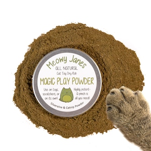 May include: A round tin container of Meowy Janes All Natural Cat Toy Dry Rub Magic Play Powder. The label features a cartoon cat and the text "Highly potent - A pinch is all you need!" The tin is surrounded by a pile of brown powder.