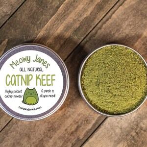 Puede incluir: Una lata redonda de metal con una etiqueta que dice "Meowy Janes All Natural Catnip Keef" con una ilustraci&oacute;n de un gato de dibujos animados. La etiqueta tambi&eacute;n dice "Highly potent catnip powder" y "A pinch is all you need!" La lata est&aacute; abierta y llena de polvo de hierba gatera verde.