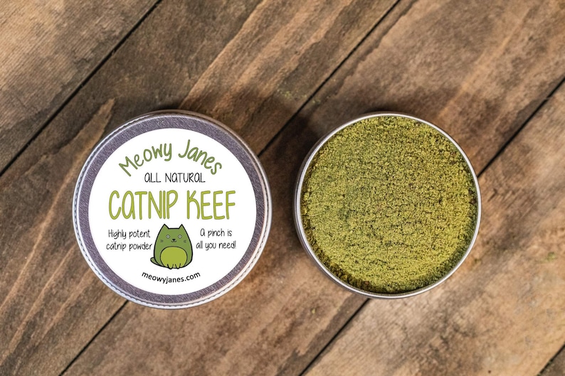 Puede incluir: Una lata redonda de metal con una etiqueta que dice "Meowy Janes All Natural Catnip Keef" con una ilustraci&oacute;n de un gato de dibujos animados. La etiqueta tambi&eacute;n dice "Highly potent catnip powder" y "A pinch is all you need!" La lata est&aacute; abierta y llena de polvo de hierba gatera verde.
