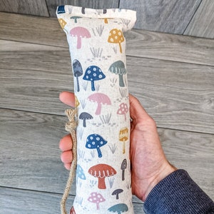 May include: A long, rectangular, white fabric toy with a brown rope handle. The fabric is covered in a pattern of colorful mushrooms with white dots. The mushrooms are in shades of pink, blue, yellow, and orange.