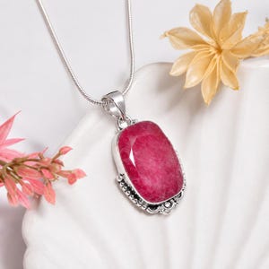 May include: A silver pendant necklace featuring a large, rectangular, faceted deep pink gemstone set in a decorative silver frame with beaded detailing along the edges, suspended from a thin, smooth silver snake chain.