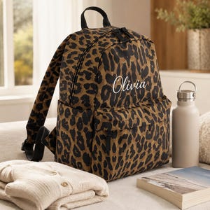 May include: A leopard print backpack featuring the name Olivia in white script lettering on the front pocket. The bag has a top handle, adjustable shoulder straps, and black zipper pulls. It is positioned next to a beige water bottle and a book.