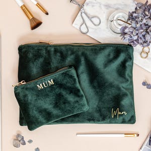 May include: A flat lay of two dark green velvet cosmetic bags with gold zips. The larger bag has the word "Mum" in gold script. A smaller bag has "MUM" in gold block letters. Makeup brushes, nail polish, and other beauty items are also present.