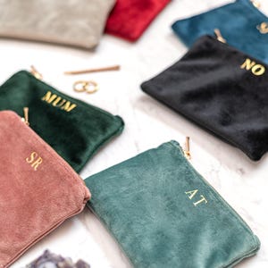 May include: A selection of velvet pouches in shades of teal, pink, and black. Each pouch features a gold zip and gold lettering, with initials or words such as "MUM" and "NO" embroidered. The pouches are displayed on a white marble surface.