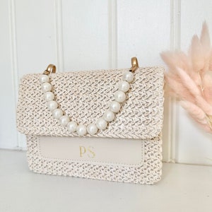 May include: A white woven straw handbag with a pearl chain strap and a cream leather flap with the initials "PS" in gold lettering.