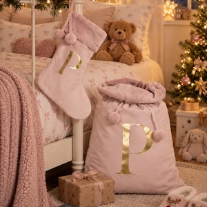 May include: A soft, light pink velvet Christmas stocking and a matching drawstring gift sack, both featuring a large, metallic gold letter P. The stocking has a plush cuff with two pom-poms, and the sack has a plush top edge with pom-poms.