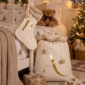 May include: A cream-colored fabric Christmas stocking with a gold letter P and a faux fur cuff, paired with a matching large drawstring gift sack featuring a gold letter A and a plush faux fur trim, both displayed in a festive bedroom setting.