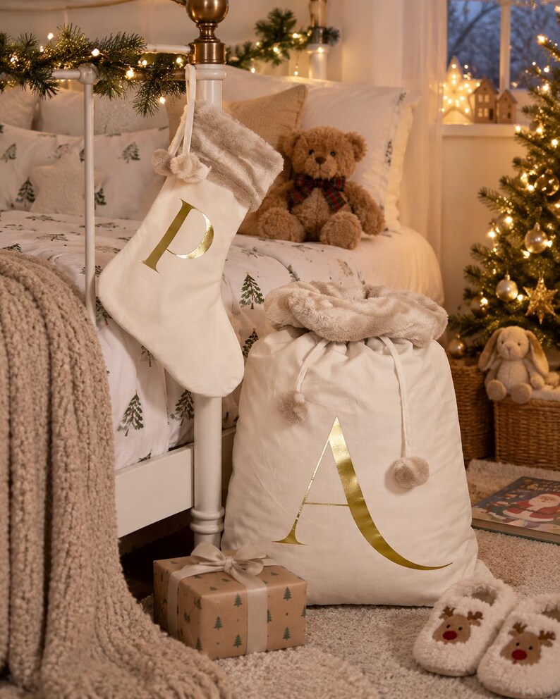 May include: A cream-colored fabric Christmas stocking with a gold letter P and a faux fur cuff, paired with a matching large drawstring gift sack featuring a gold letter A and a plush faux fur trim, both displayed in a festive bedroom setting.