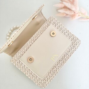May include: A cream-colored woven straw handbag with a flap closure and a pearl chain strap. The flap has a cream-colored leather lining with the initials "PS" embossed in gold.