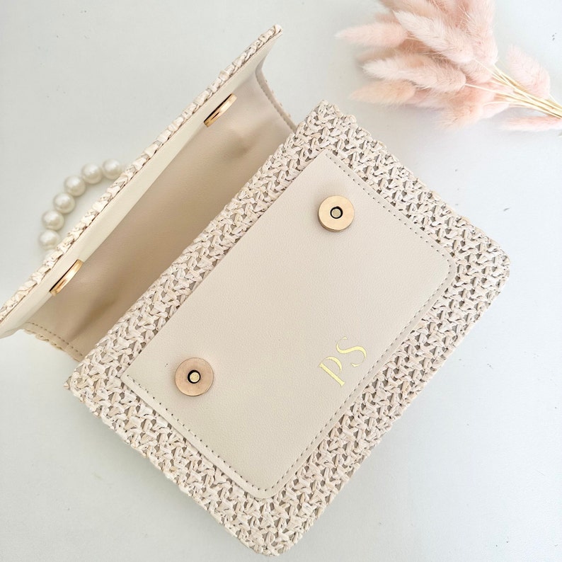 May include: A cream-colored woven straw handbag with a flap closure and a pearl chain strap. The flap has a cream-colored leather lining with the initials "PS" embossed in gold.