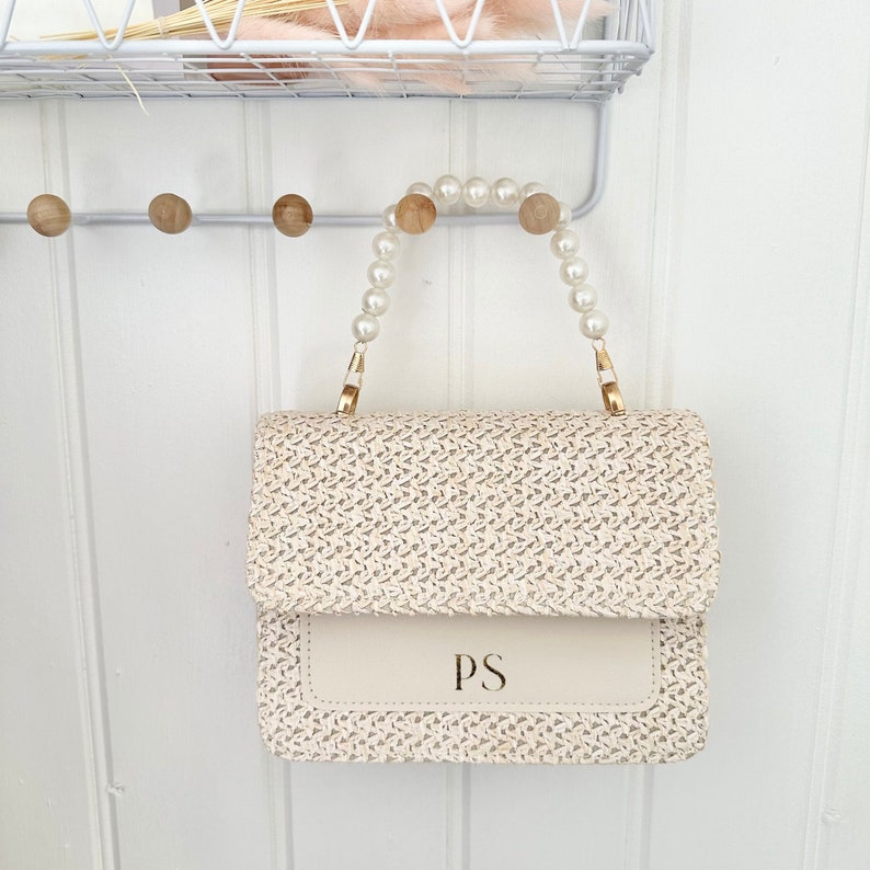 May include: A cream-colored woven handbag with a pearl and gold chain strap. The bag has a flap with the initials "PS" in gold lettering.