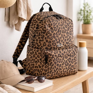 May include: A leopard print backpack featuring black zippers, a top handle, and the white embroidered initials RS on the front pocket. The bag has a main compartment and a smaller front pocket, both secured with zippers, and adjustable shoulder straps.