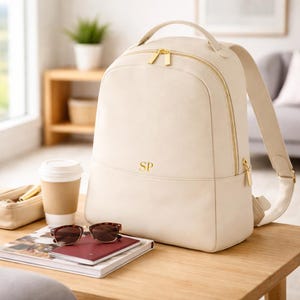 May include: Cream-colored backpack with gold-tone zippers and hardware. The backpack has a top handle and adjustable shoulder straps. The initials "SP" are displayed in gold on the front. Sunglasses, a passport, and a coffee cup sit on a wooden table.
