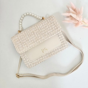 May include: A white woven straw handbag with a pearl handle and a long strap. The bag has a white leather flap with the initials "PS" in gold lettering.