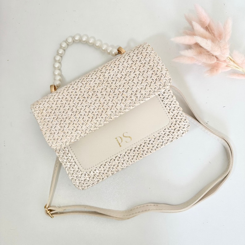 May include: A white woven straw handbag with a pearl handle and a long strap. The bag has a white leather flap with the initials "PS" in gold lettering.