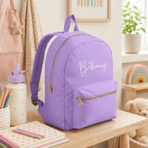 May include: A lavender backpack featuring gold-tone zippers and the name Bethany printed in white script on the front pocket. The bag includes a top handle and adjustable shoulder straps, with a main compartment and a smaller front storage pocket.