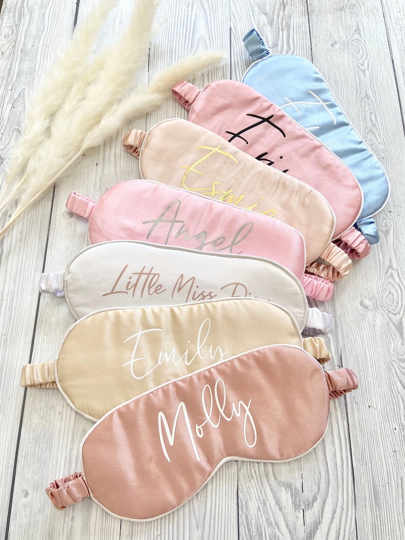 May include: Six pink, beige, and blue satin sleep masks with white piping. Each mask has a different name embroidered on it in gold or white thread. The names are: "Molly", "Emily", "Little Miss D", "Angel", "Essie", and "Hailey".