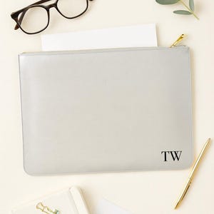 May include: A light gray leather document case with a gold zipper, the initials "TW" are printed in black on the lower right corner. A gold pen, glasses, and a notebook are also visible.