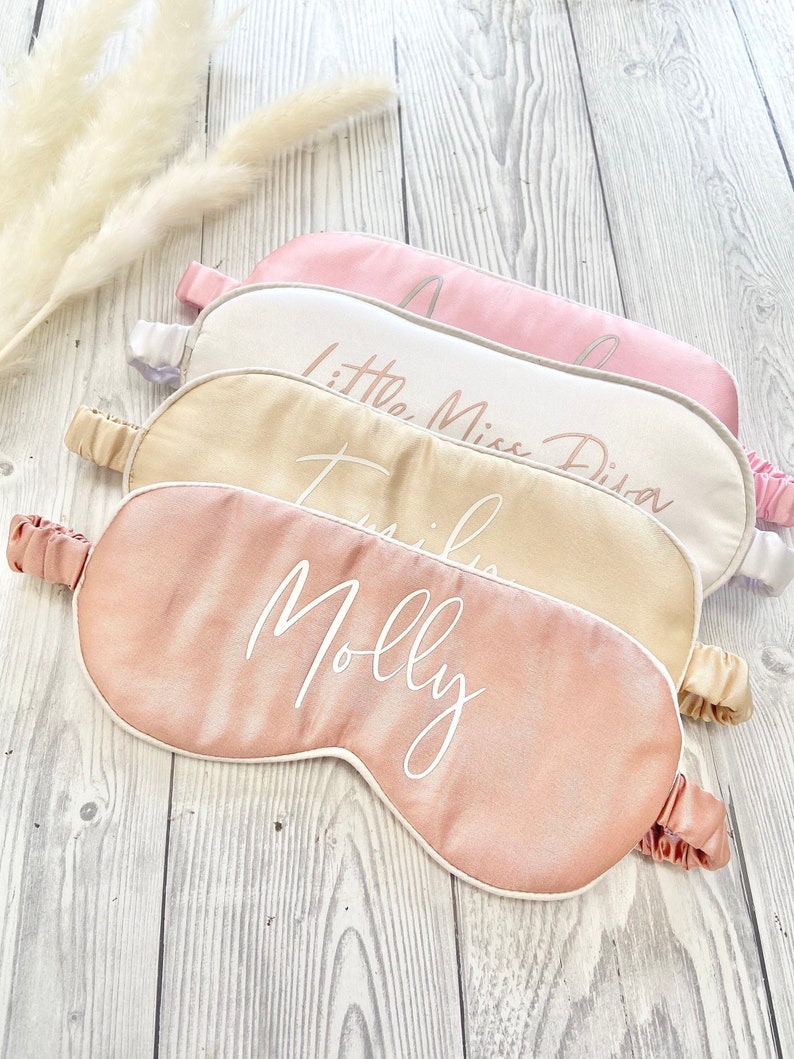 May include: Four satin sleep masks with white piping and personalized names in white script. The masks are pink, white, and beige.
