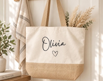 May include: A cream-colored canvas tote bag with a tan burlap base and two matching burlap handles. The front of the bag features the name Olivia printed in black script lettering above a small, simple black heart outline graphic.