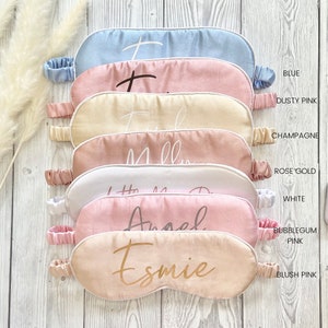 May include: Six satin sleep masks in various colors, including blue, dusty pink, champagne, rose gold, white, and blush pink. Each mask has a different name embroidered on it in gold lettering: "I", "Holly", "Millie", "Little Miss D", "Angel", and "Esmie".