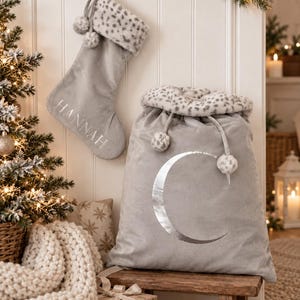 May include: A light gray velvet Christmas stocking with the name HANNAH embroidered in silver, featuring a leopard print faux fur cuff and pom-poms. Beside it sits a matching large gray velvet gift sack with a silver crescent moon and leopard trim.