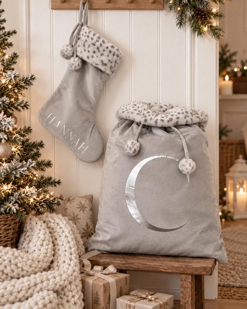 May include: A light gray velvet Christmas stocking with the name HANNAH embroidered in silver, featuring a leopard print faux fur cuff and pom-poms. Beside it sits a matching large gray velvet gift sack with a silver crescent moon and leopard trim.