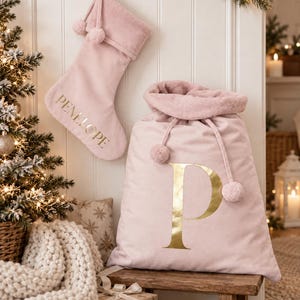 May include: A soft, light pink velvet Christmas stocking with the name PENELOPE in gold lettering and a matching light pink velvet Santa sack featuring a large gold letter P. Both items include drawstring closures with decorative pom-pom accents.